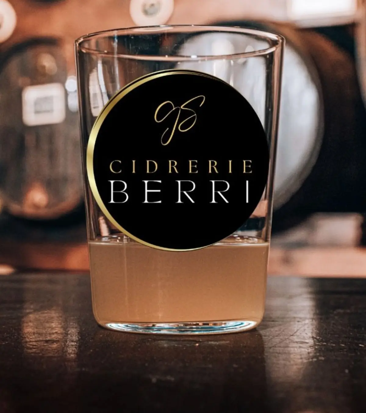 Verre de Sagarno marqué du logo Cidrerie Berri, posé devant les fûts de chêne du chai